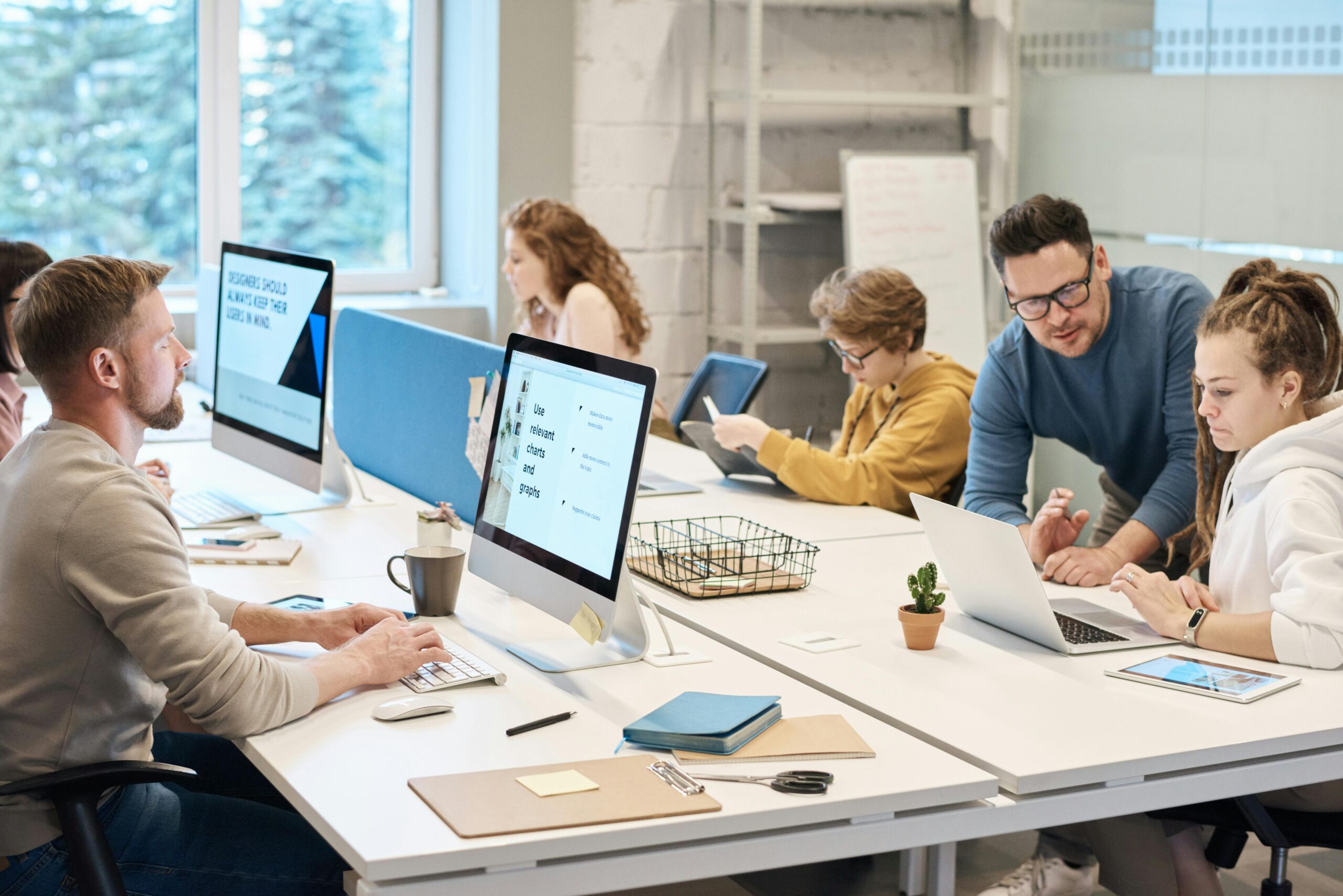Modern UI/UX design agency workspace with designers working on multiple monitors displaying Figma interfaces and wireframes